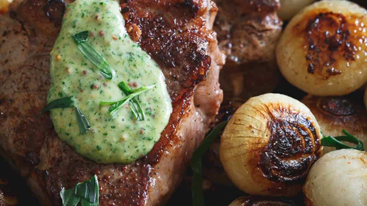 Grilled steak topped with herb butter, served alongside charred whole onions and garnished with fresh herbs.