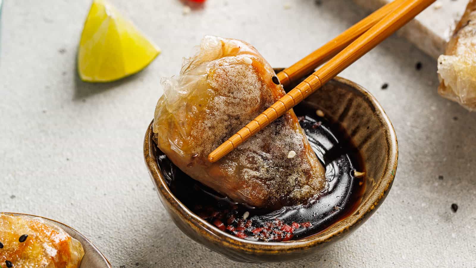 A piece of fried spring roll held by chopsticks is dipped into a bowl of soy sauce, with a lime wedge in the background.