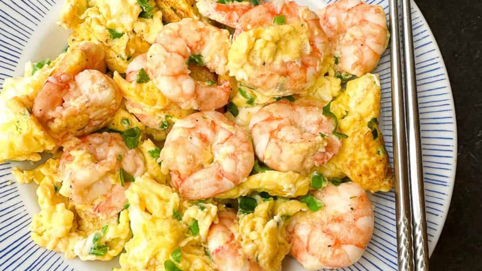 A plate of scrambled eggs mixed with cooked shrimp and chopped green onions, served with a pair of metal chopsticks.