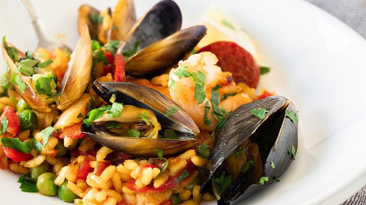 A plate of seafood paella with rice, mussels, shrimp, peas, and chopped herbs, garnished with a lemon wedge.