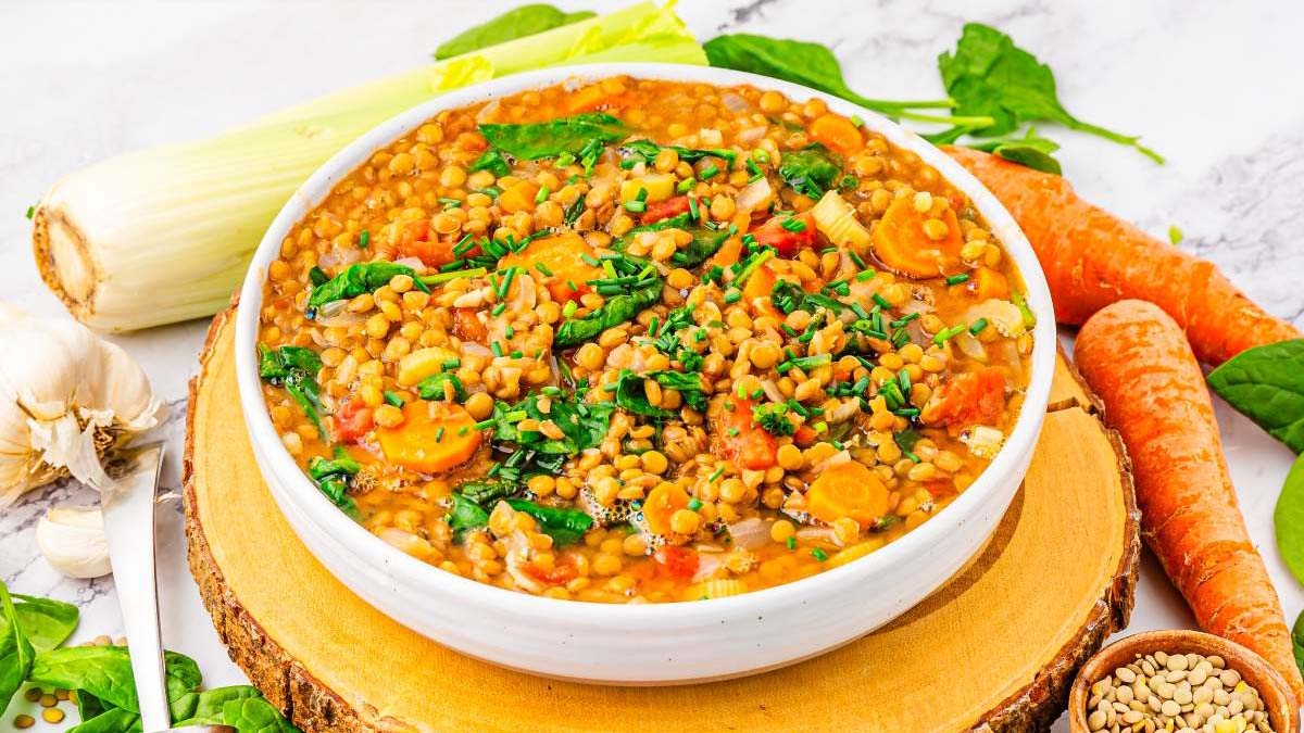 A bowl of lentil soup with sliced carrots, spinach, and chopped herbs, surrounded by fresh vegetables on a wooden board.