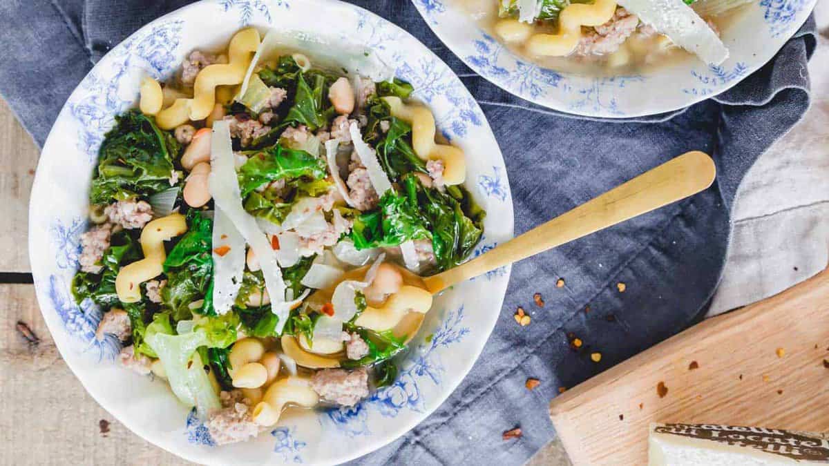 A bowl of soup with curly pasta, white beans, kale, ground meat, and cheese shavings, served with a gold spoon on a blue-patterned plate.