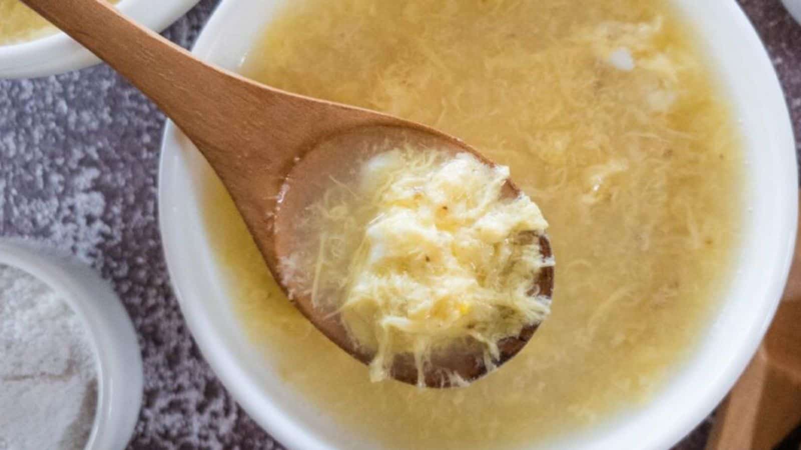 A wooden spoon holding a portion of egg drop soup over a bowl filled with the same soup.