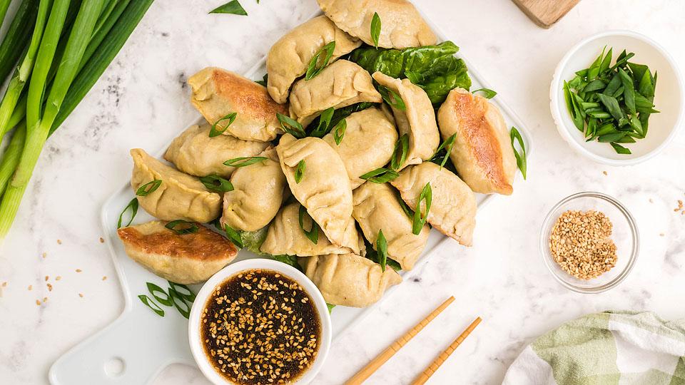A pile of cooked dumplings garnished with sliced green onions, served with a bowl of dipping sauce, sesame seeds, and extra onions on a marble surface.