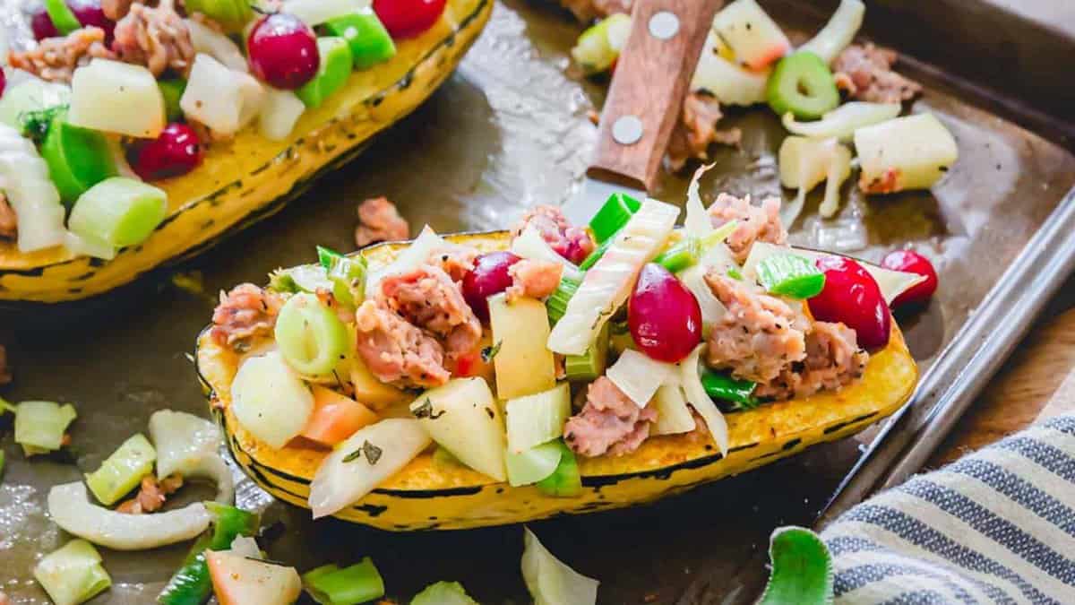 A baked squash half filled with a mixture of cooked sausage, chopped apples, celery, scallions, and cranberries, on a baking sheet.