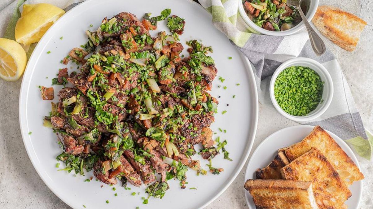A white plate of grilled meat topped with chopped herbs and onions, served with toasted bread, lemon wedges, and bowls of green seasoning and extra sauce.