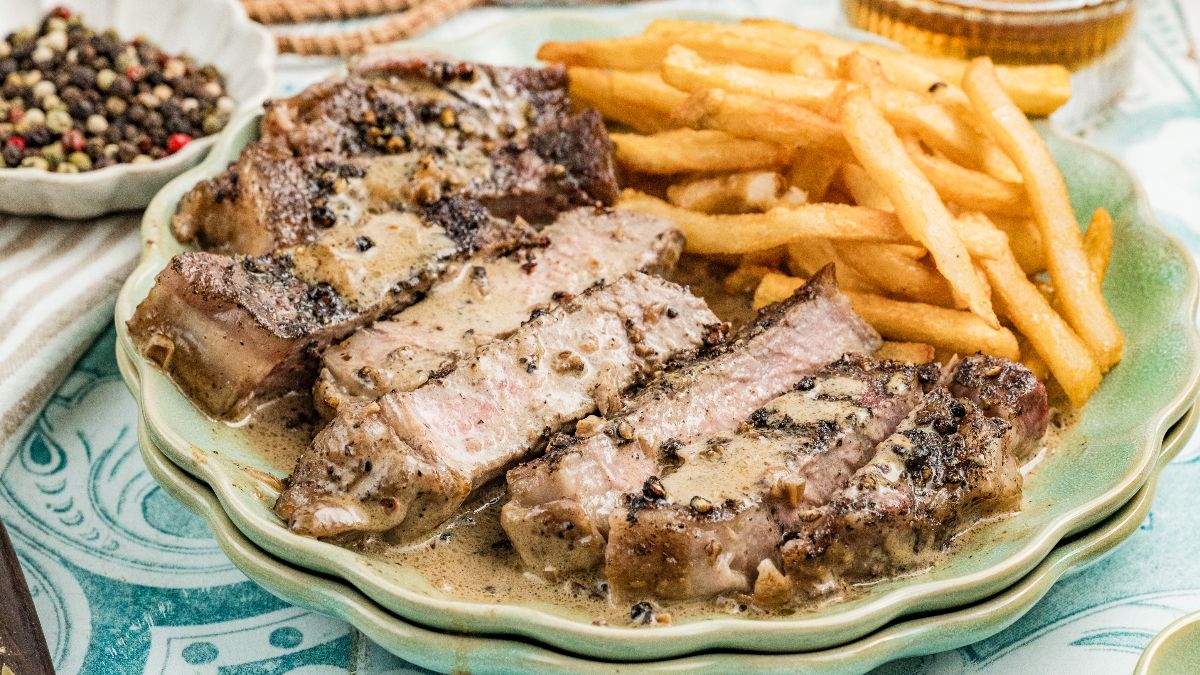 A plate of sliced steak with peppercorn sauce, served with a side of French fries.