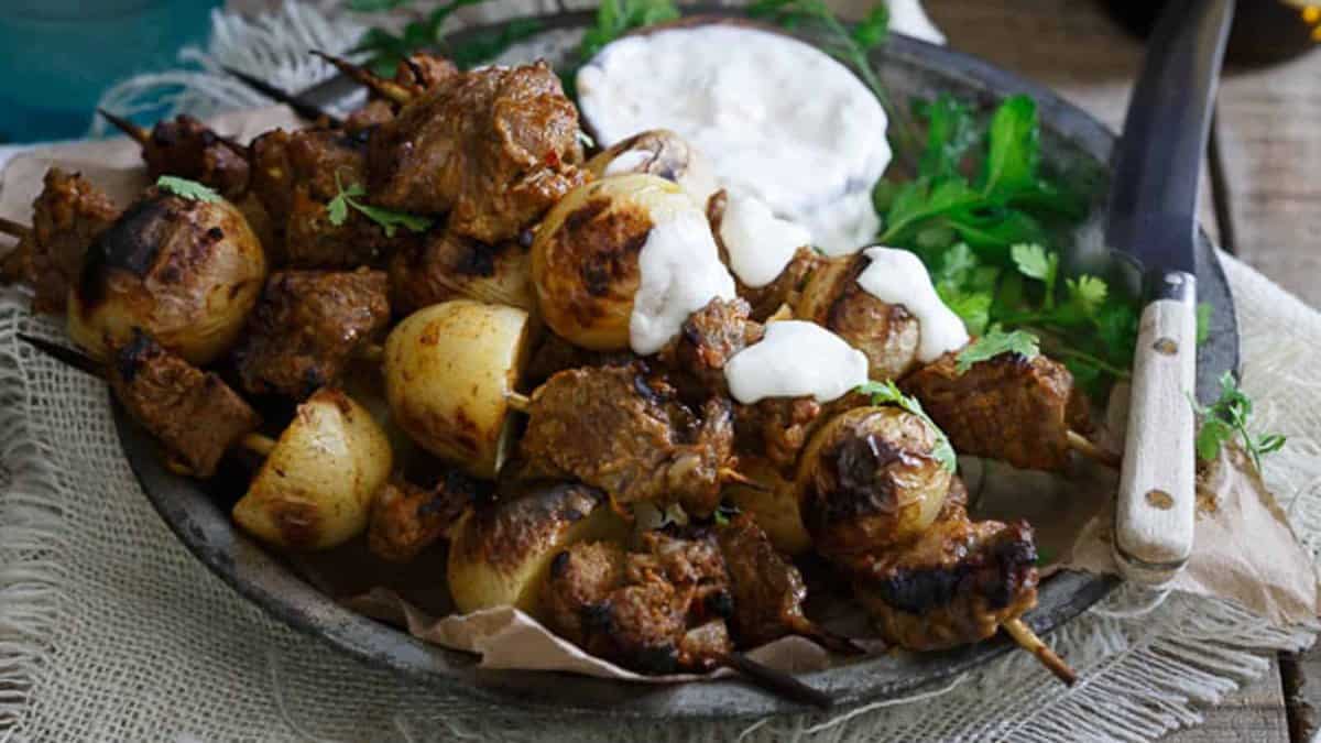 Plate of grilled meat and onion skewers garnished with herbs, served with a side of white dipping sauce and a knife on a rustic cloth.