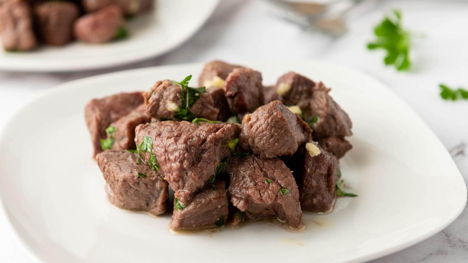 A white plate with cooked beef cubes garnished with chopped herbs, with another plate of beef and some parsley in the background.