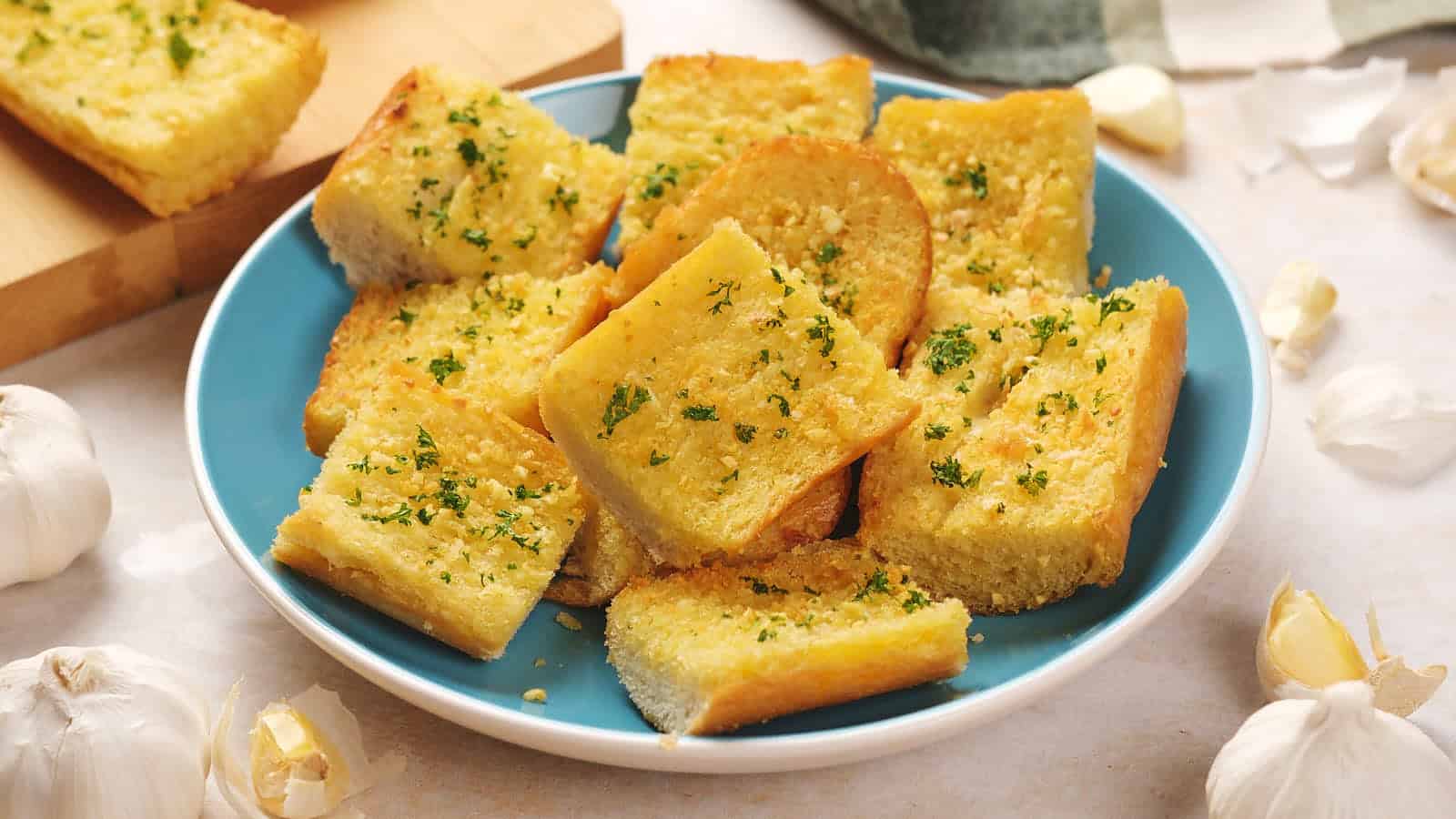 A blue plate filled with slices of garlic bread garnished with chopped parsley, surrounded by garlic cloves on a light surface.