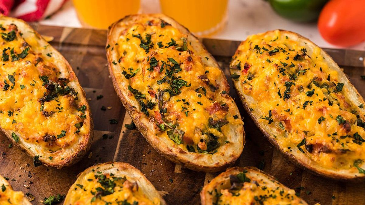 Baked potato skins filled with melted cheese, herbs, and diced vegetables, arranged on a wooden board with drinks in the background.