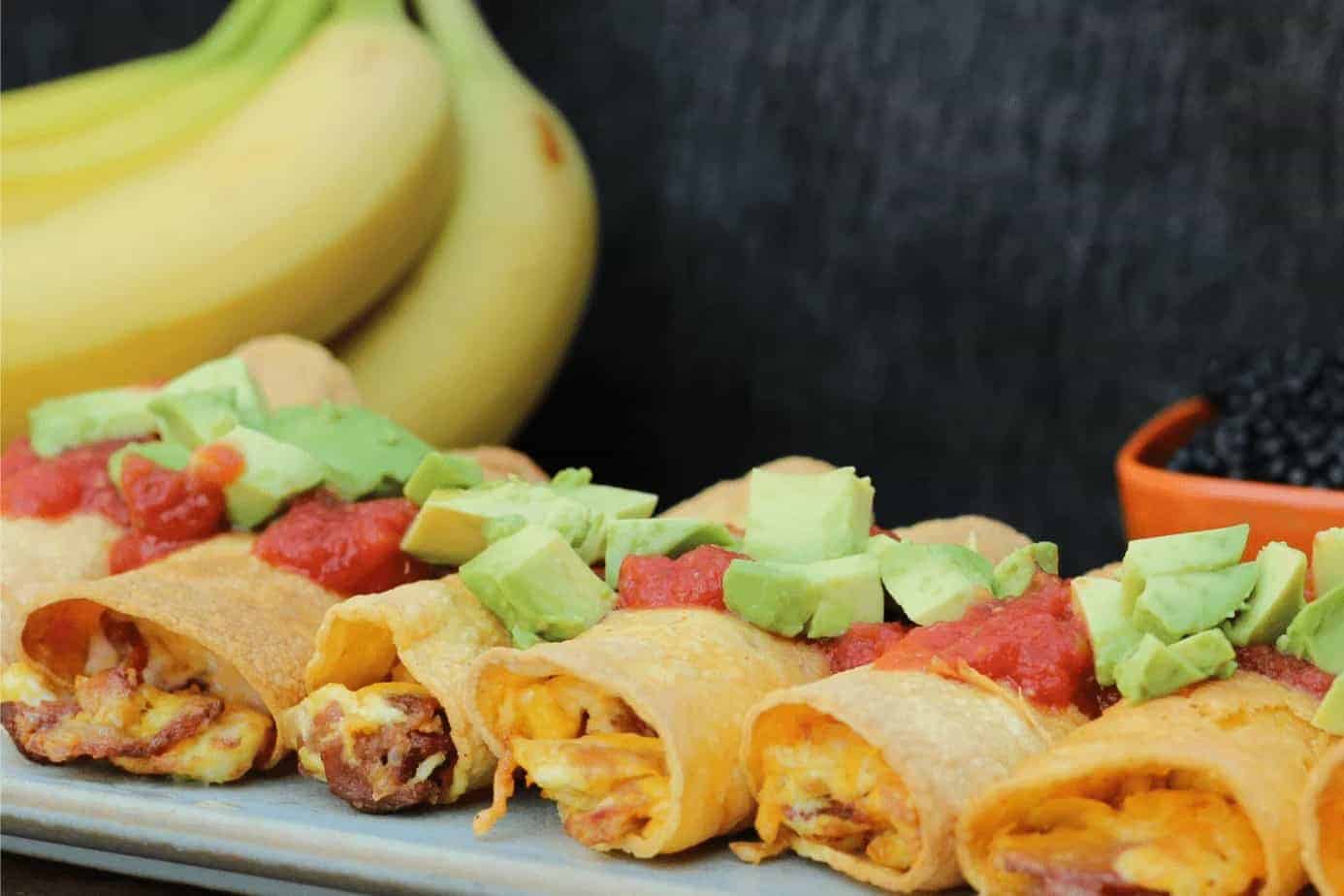 A row of breakfast tacos topped with diced avocado and salsa is displayed on a plate, with bananas and a bowl of blackberries in the background.
