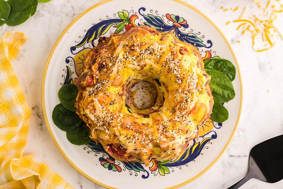 A savory bundt-shaped bread topped with cheese and seeds sits on a decorative plate with spinach leaves underneath.