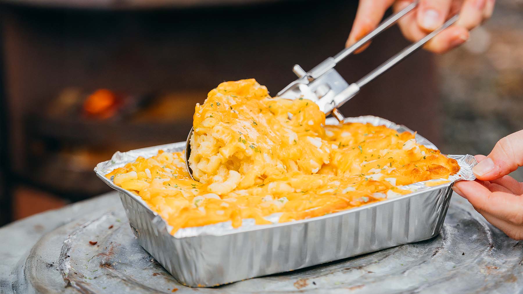 A hand scoops a serving of baked macaroni and cheese from a foil tray with metal tongs.