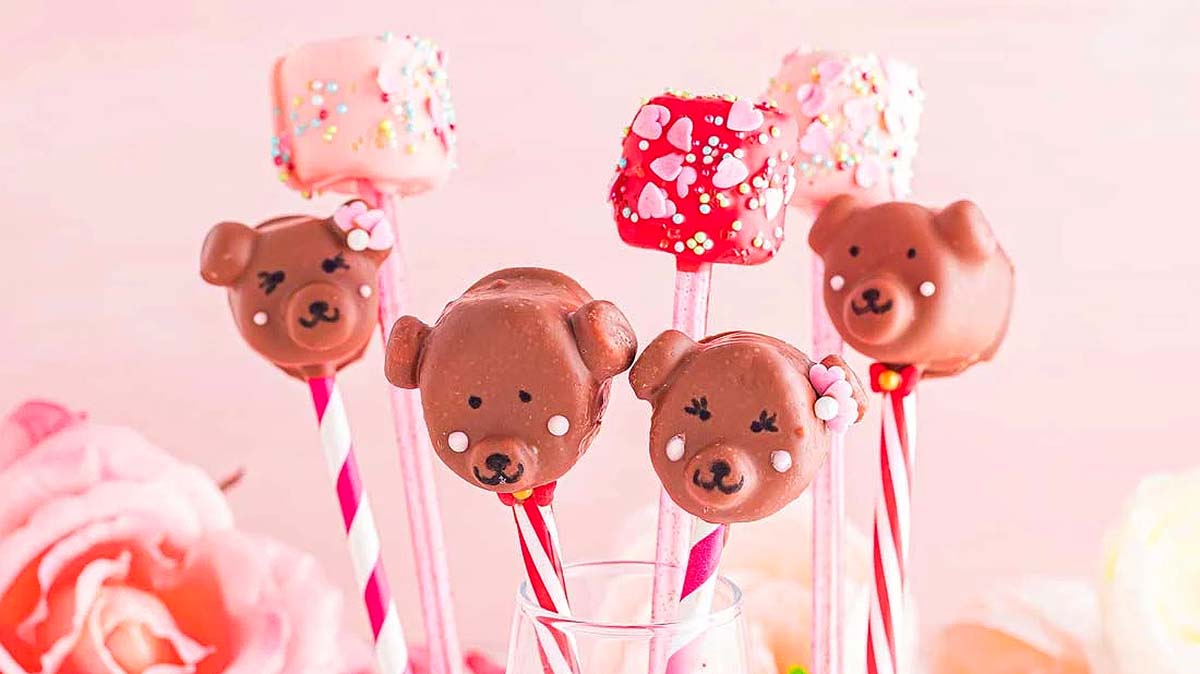 Colorful cake pops decorated as bear faces and sprinkled marshmallows on sticks, displayed in a glass with a pink floral background.