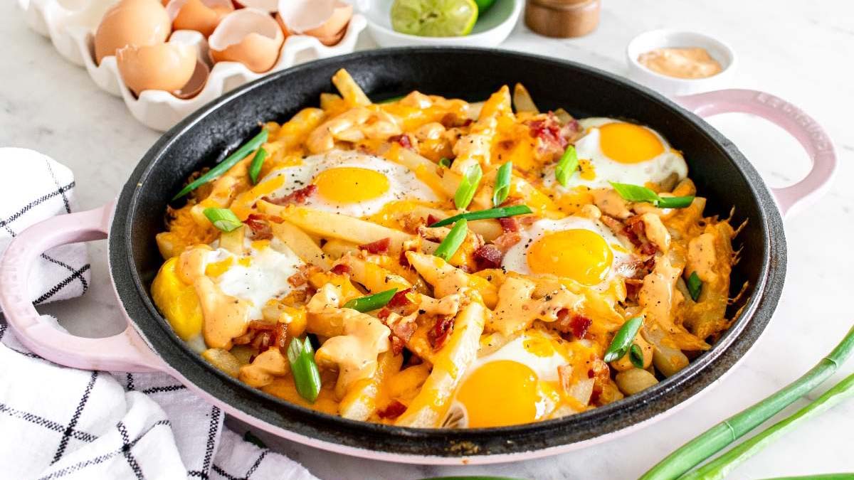 A skillet filled with fries, melted cheese, bacon, and four sunny-side-up eggs, garnished with chopped green onions. Eggshells and ingredients are visible in the background.