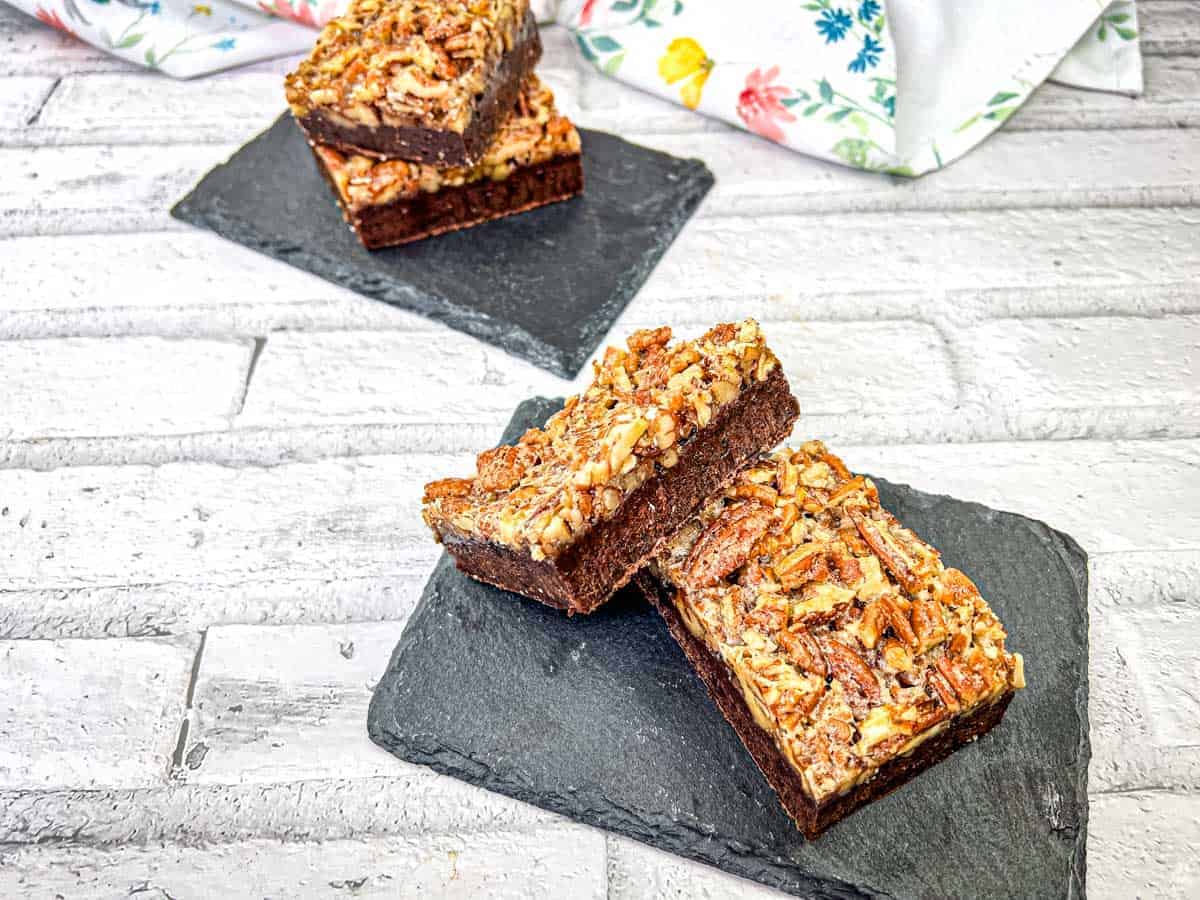 Two pecan-topped brownies are placed on a black slate, with two more brownies stacked in the background on another slate, set on a white brick surface.