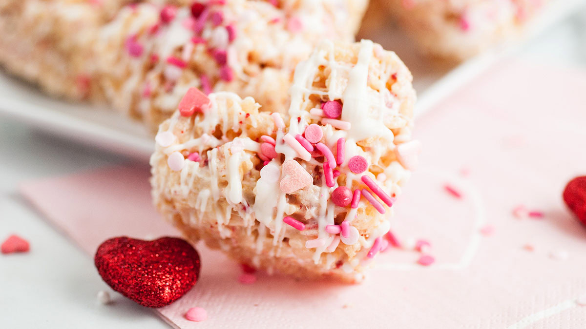 Heart-shaped rice cereal treat topped with white icing and pink, red, and white sprinkles, placed on a pink napkin with red glitter hearts nearby.