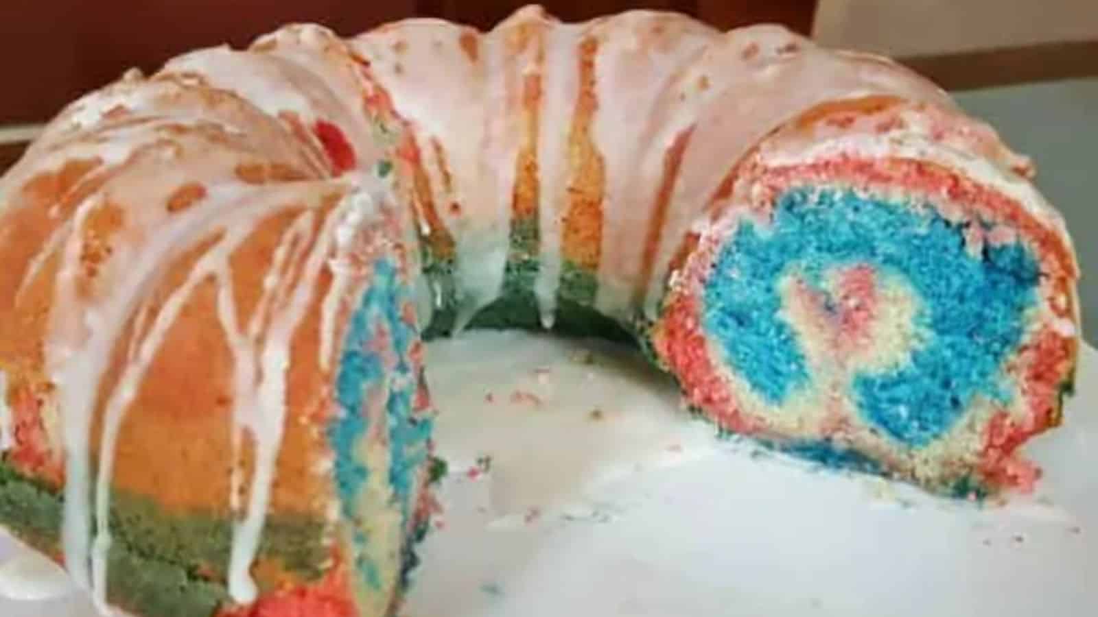 A bundt cake with red, blue, and green layers, covered in white icing, with a large slice cut to show the colorful interior.