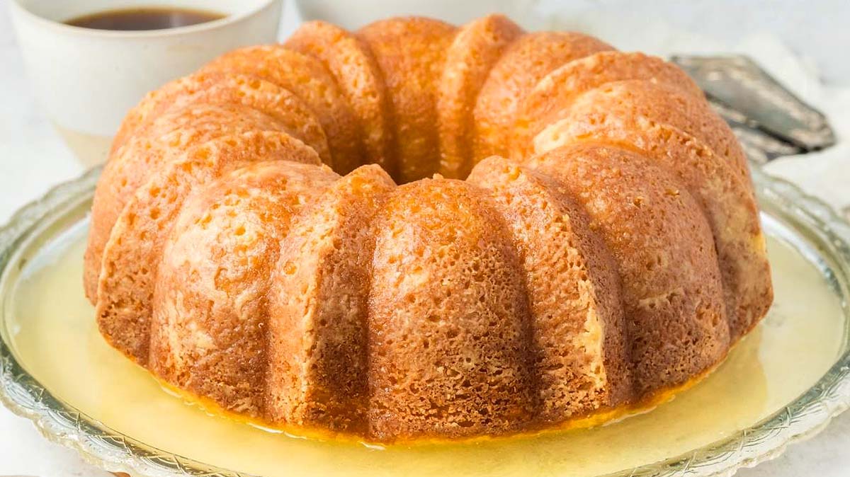 A glazed bundt cake sits on a glass plate, with a bowl of liquid and utensils in the background.