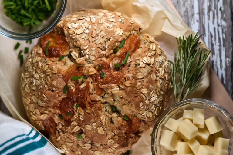 A round loaf of oat-topped bread sits on parchment paper, surrounded by fresh chives, rosemary, and a bowl of cubed cheese.