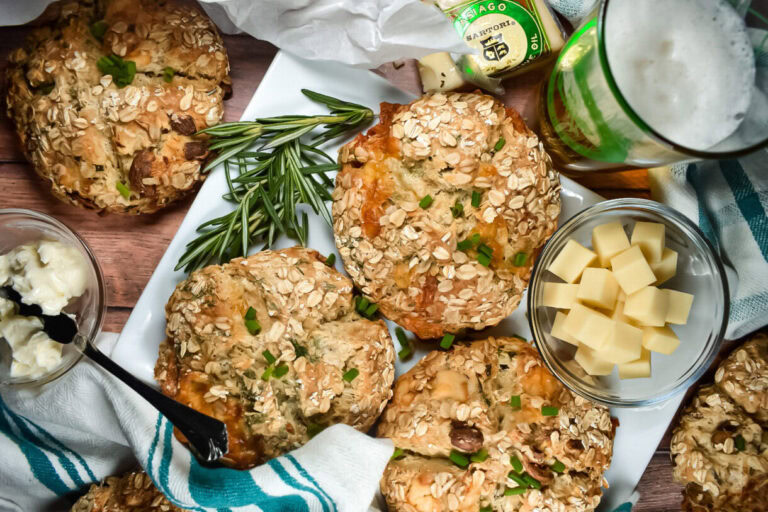 Round oat-topped bread rolls on a tray, garnished with rosemary, next to a bowl of cheese cubes, a glass of beer, butter, and a striped towel.