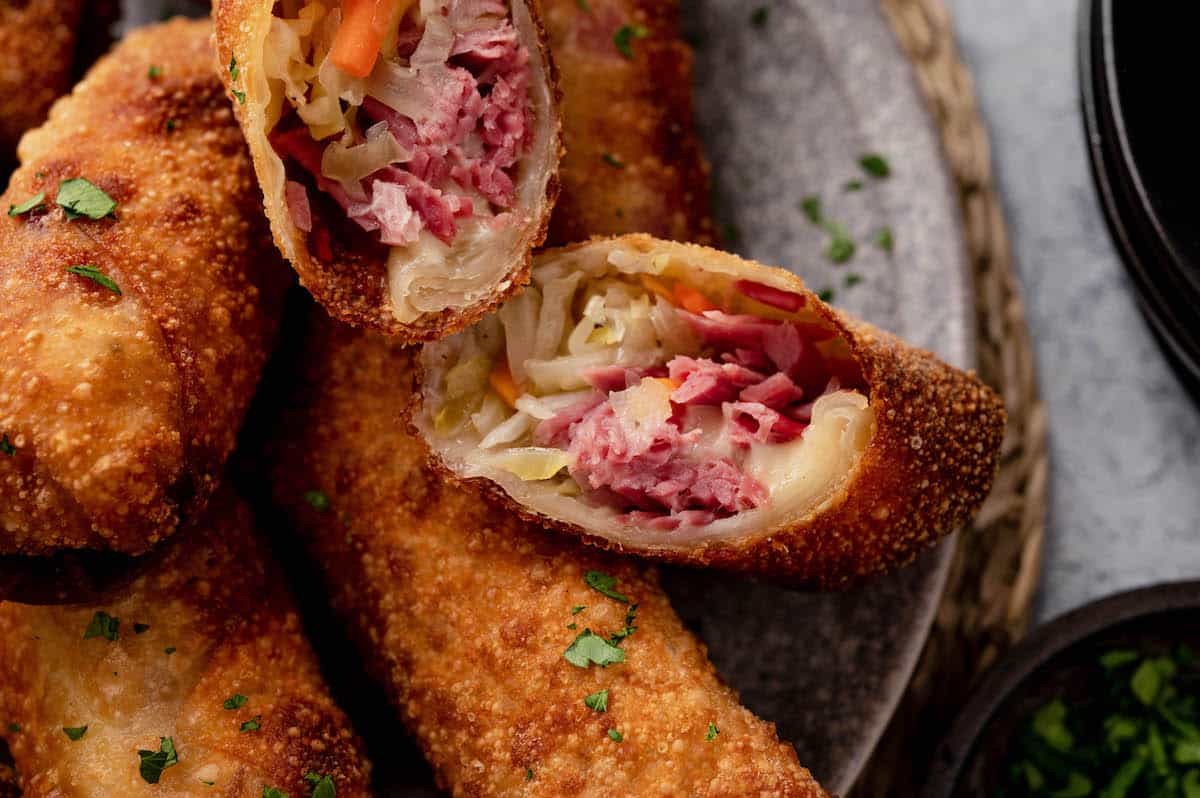 A close-up of crispy egg rolls sliced open, showing a filling of corned beef, sauerkraut, and melted cheese on a gray plate.
