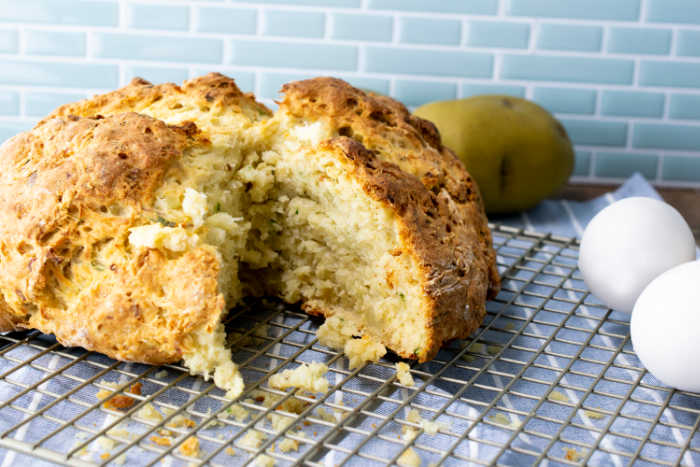 A round loaf of bread with a slice removed sits on a wire cooling rack, with two eggs and a potato in the background.