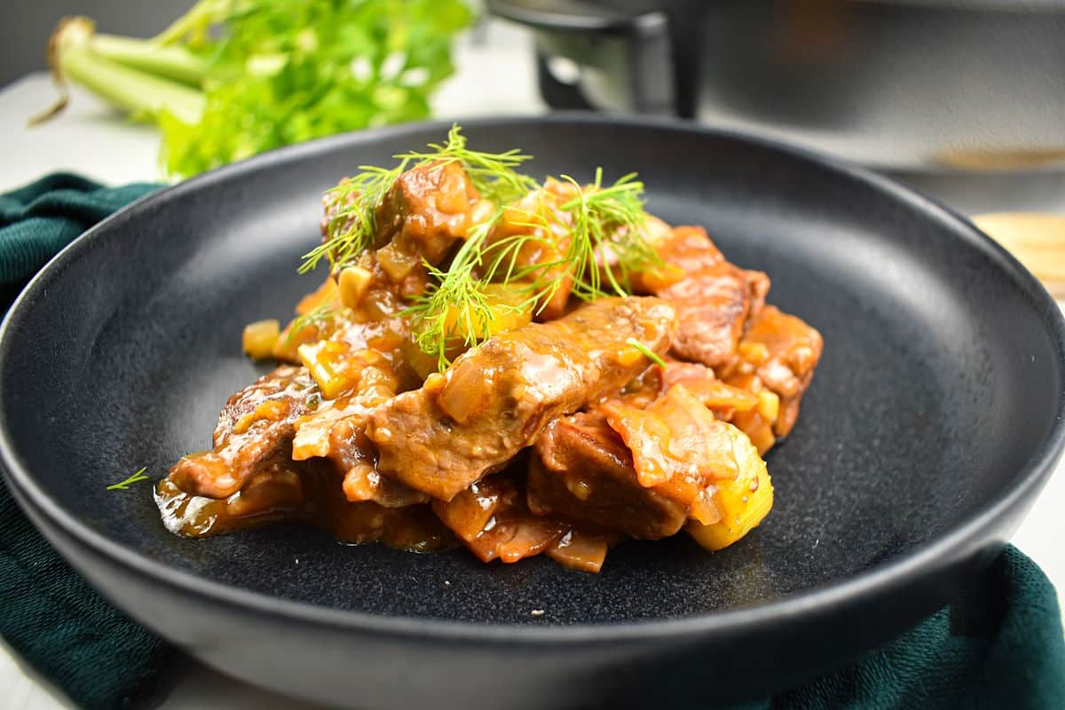 A black plate with stewed beef pieces in a brown sauce, garnished with fresh dill, and celery in the background.