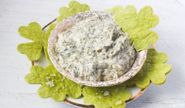 A bowl of creamy spinach dip is placed on a plate surrounded by green, leaf-shaped crackers.