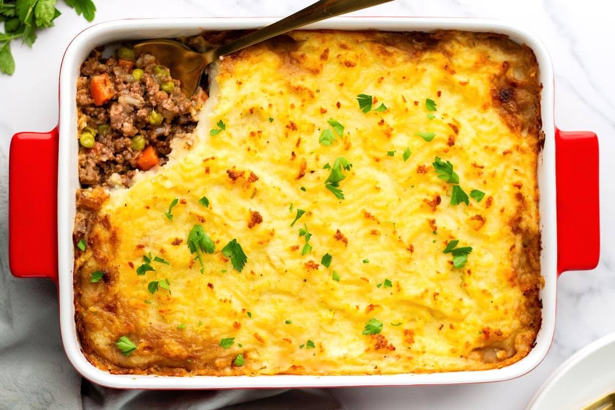 A baked casserole dish of shepherd’s pie with a portion removed, showing layers of ground meat, vegetables, and mashed potato topping, garnished with chopped parsley.