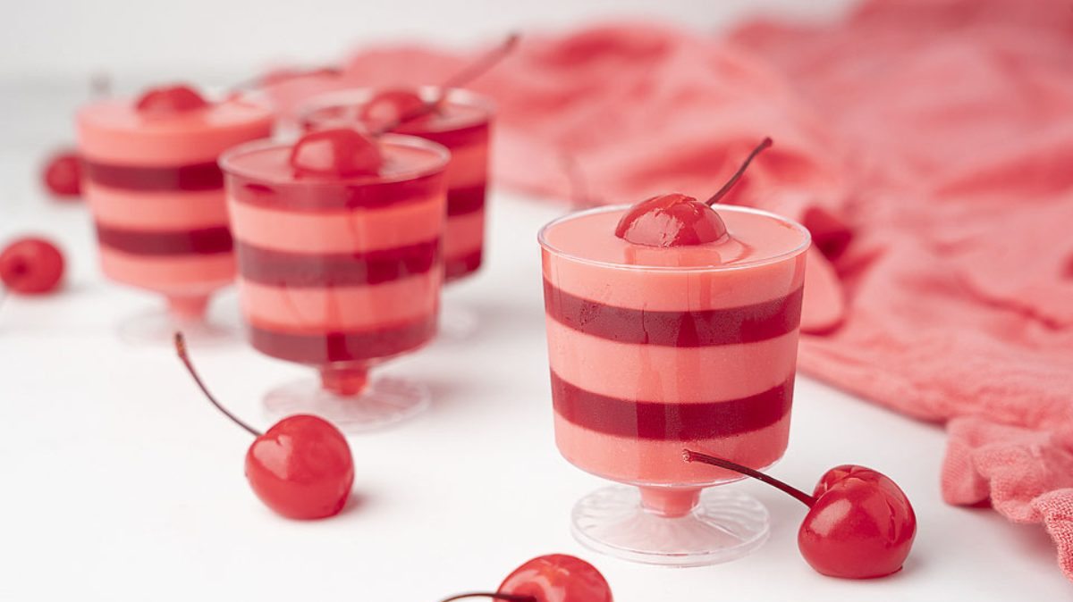 Four layered cherry and chocolate mousse desserts in clear cups, each topped with a cherry, are displayed on a white surface with loose cherries and a pink cloth in the background.