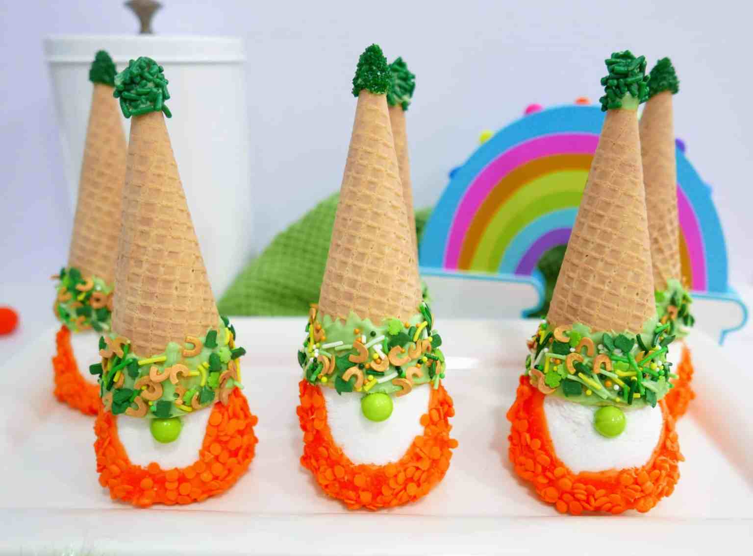 Four marshmallow treats decorated as gnomes with orange sprinkles, green candy, and ice cream cone hats, arranged on a white tray with a rainbow decoration in the background.