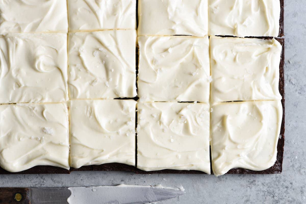 A batch of frosted brownies cut into squares on a gray surface, with a knife resting nearby.