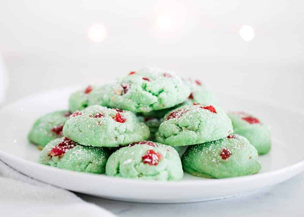 A plate with a pile of green cookies containing red bits, lightly dusted with powdered sugar, on a white surface.