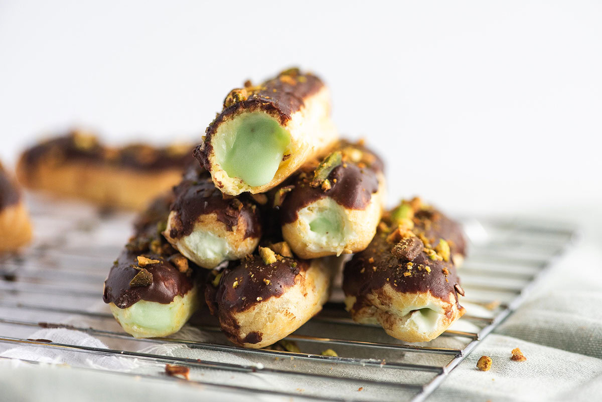A stack of chocolate-glazed éclairs filled with green cream and sprinkled with crushed pistachios, displayed on a cooling rack.