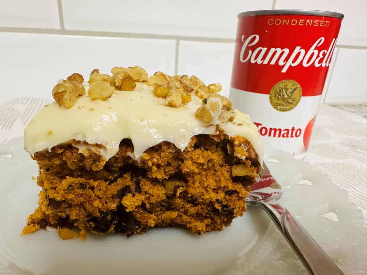 A slice of frosted carrot cake topped with walnuts on a plate with a fork, next to a can of Campbellโs condensed tomato soup.