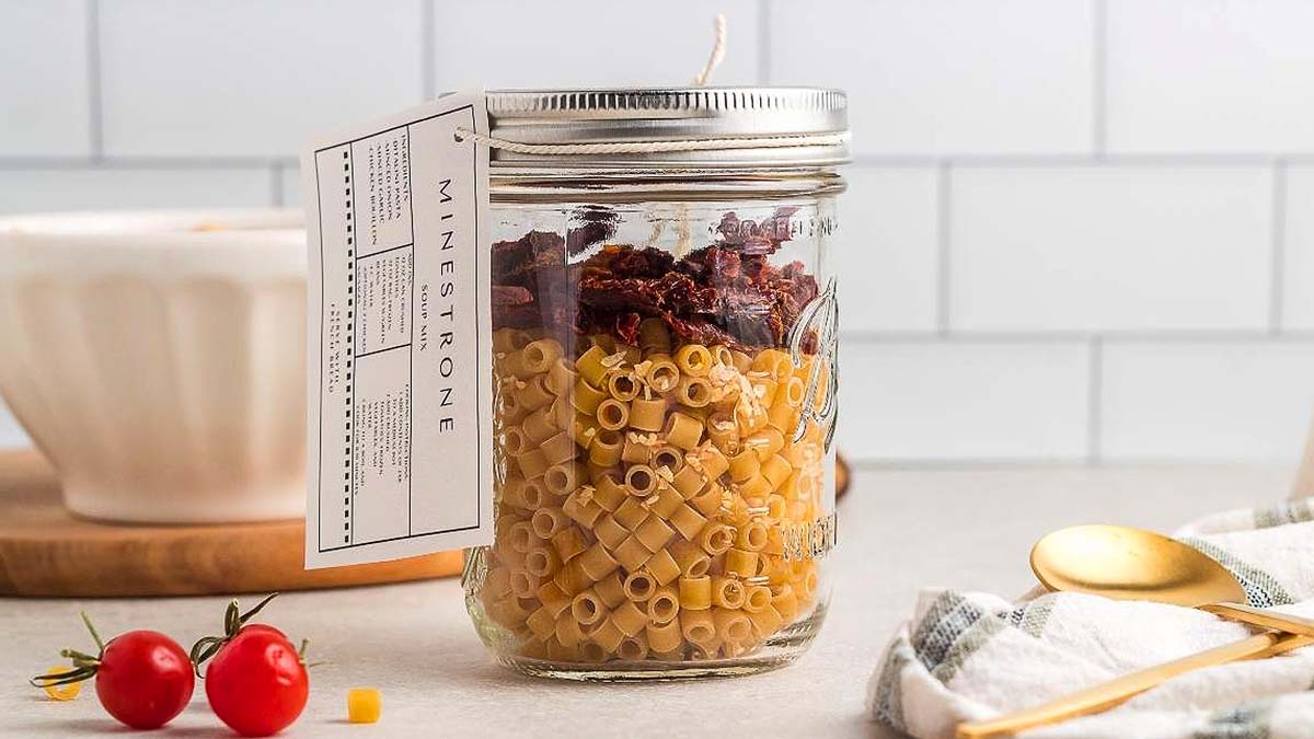 A mason jar filled with dry pasta and sun-dried tomatoes, labeled Minestrone, sits on a kitchen counter beside cherry tomatoes, a spoon, and a towel.