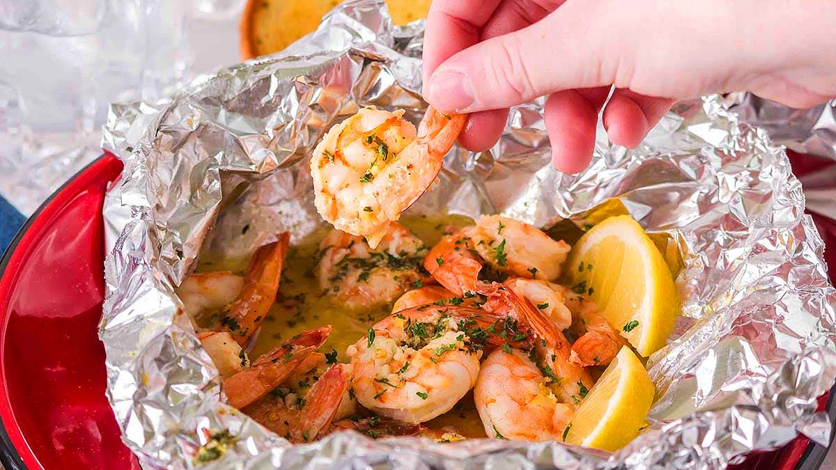 A hand holds a cooked shrimp over a foil packet filled with shrimp, herbs, and lemon wedges.
