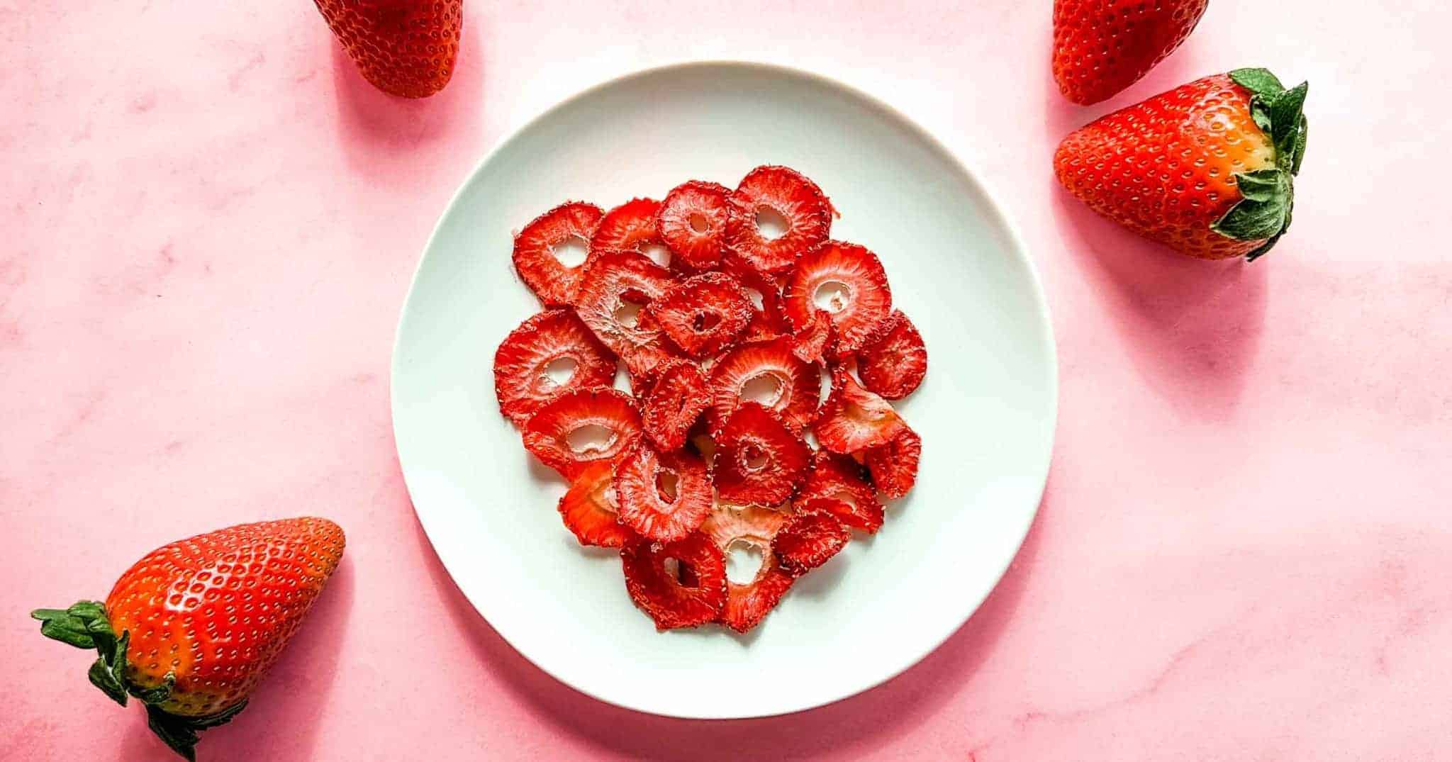 A white plate with dried strawberry slices on a pink surface, surrounded by four whole strawberries.