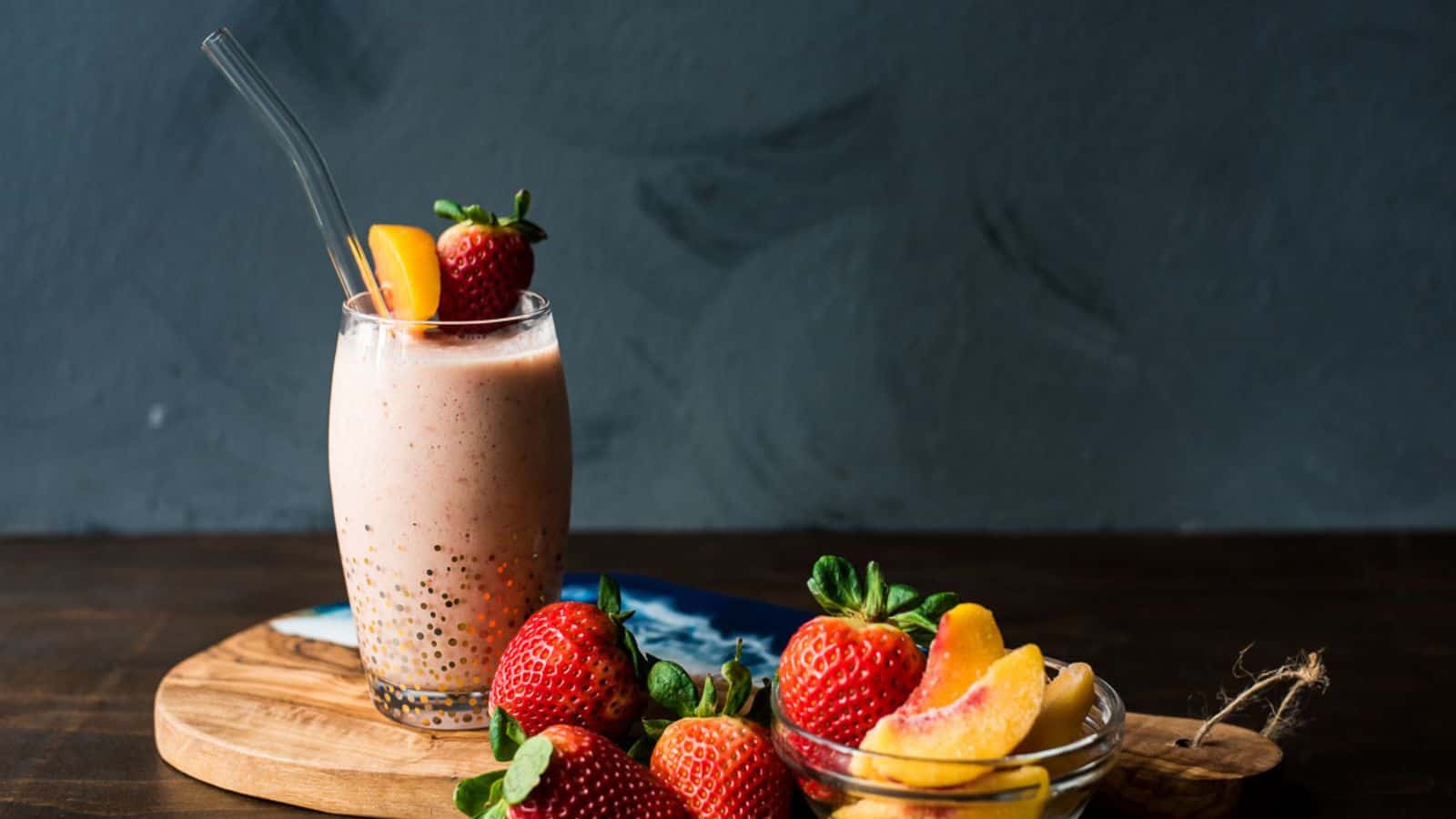 A glass of strawberry smoothie with chia seeds and a peach slice, next to fresh strawberries and sliced peaches on a wooden board.