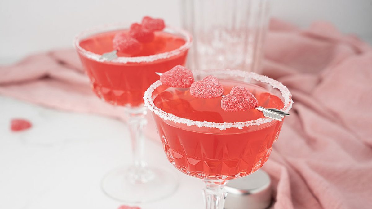 Two pink cocktails in vintage glasses with sugar rims, garnished with pink gummy candies on skewers, set on a light pink cloth.