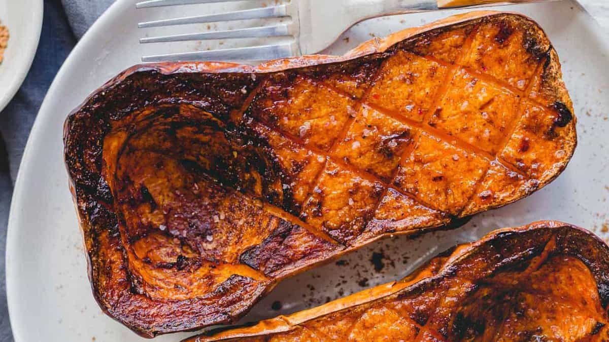 Close-up of two roasted butternut squash halves on a white plate, scored with a crosshatch pattern and seasoned, with a fork in the background.