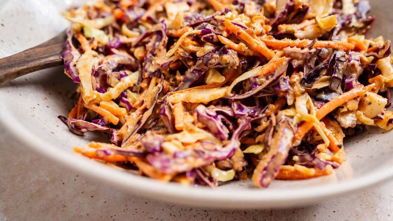 A close-up of a bowl filled with coleslaw made of shredded red cabbage, carrots, and creamy dressing, with a wooden spoon resting inside.