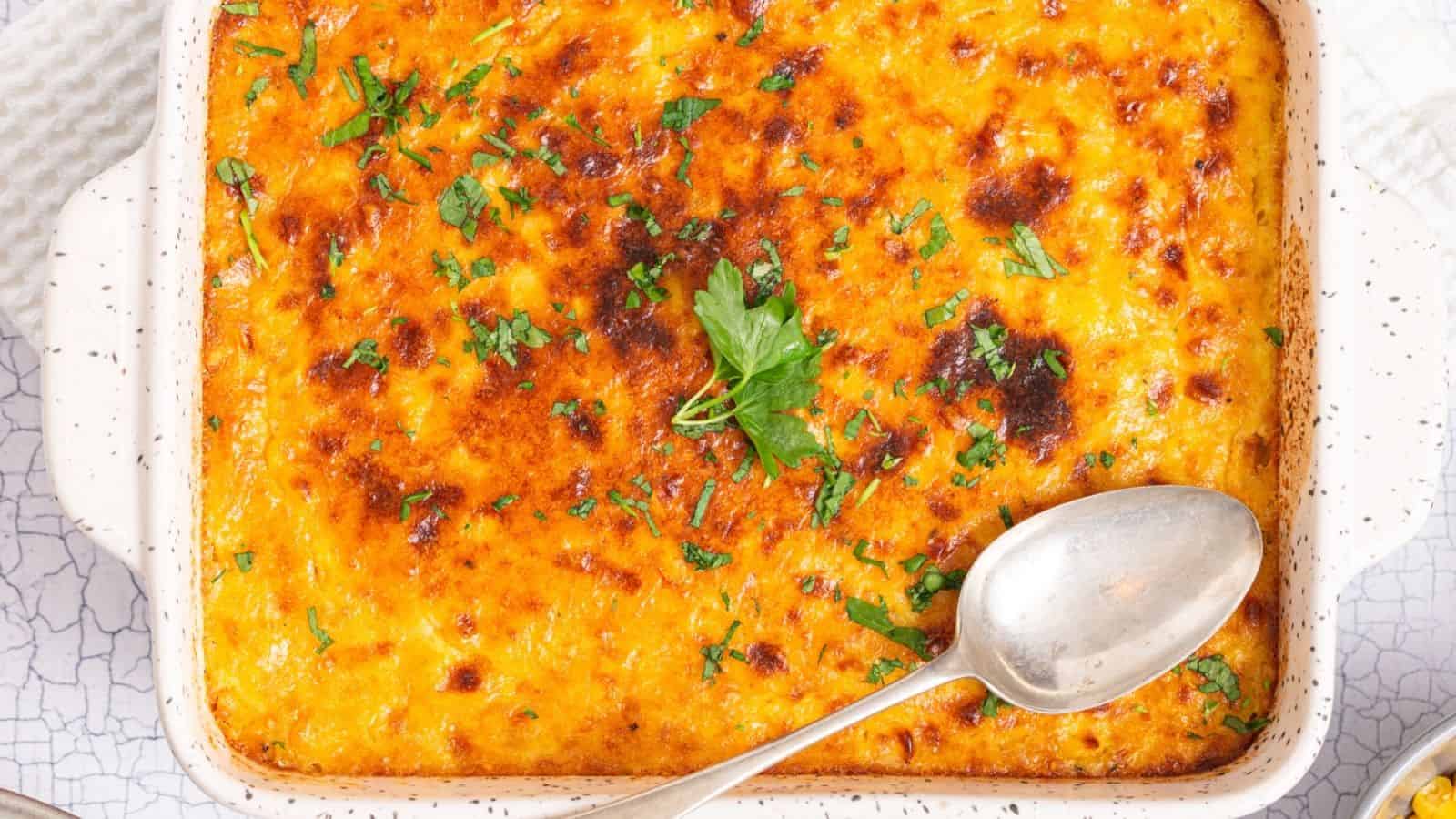 A baked casserole with a golden, slightly browned cheese topping, garnished with chopped herbs, in a white square dish with a serving spoon.