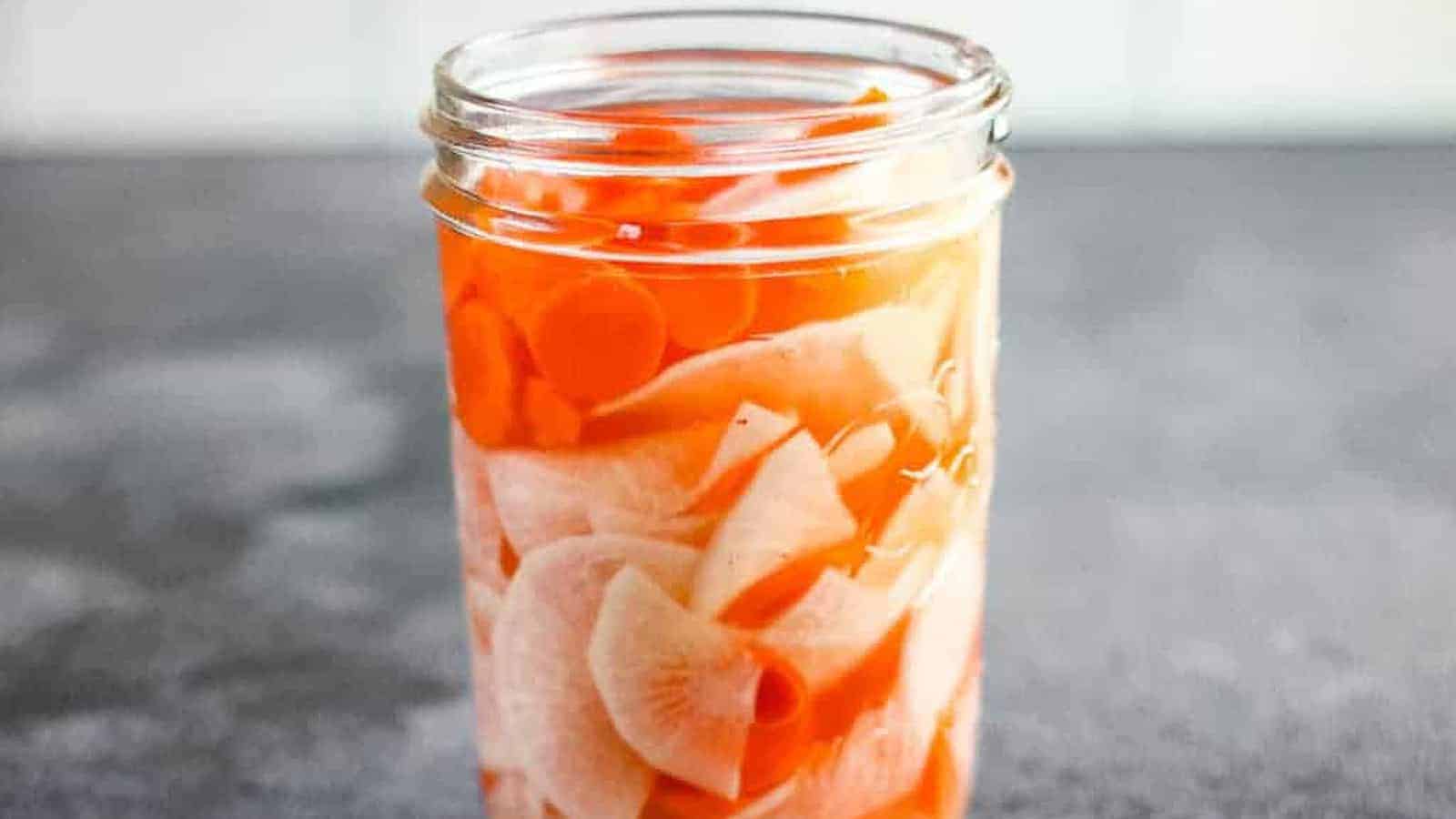 A glass jar filled with pickled white radish and orange carrot slices in brine, placed on a gray surface.