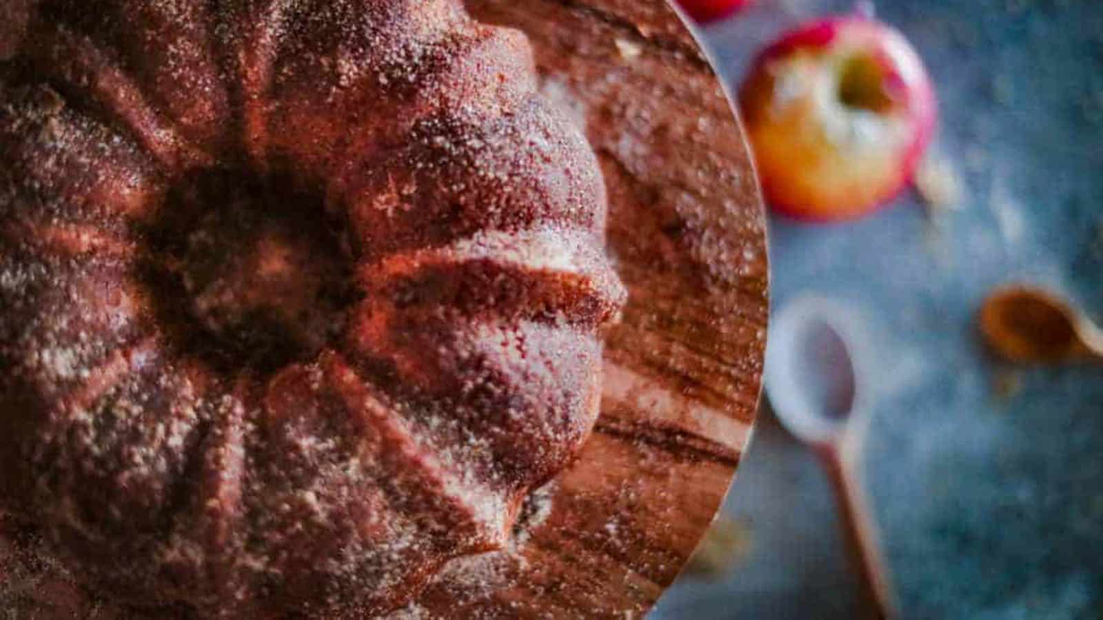 A bundt cake with a sugar dusting sits on a wooden surface, with apples and a wooden spoon blurred in the background.
