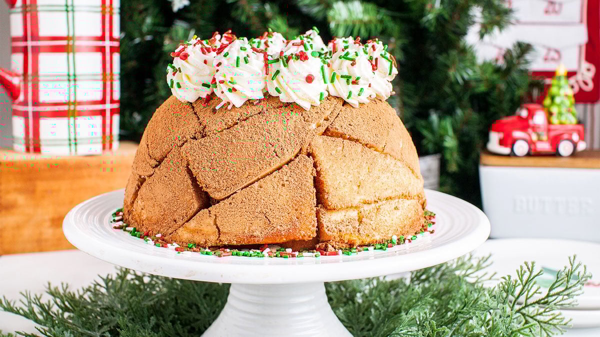 A dome-shaped Christmas cake topped with whipped cream and red, green, and white sprinkles sits on a white cake stand in front of a decorated Christmas tree.