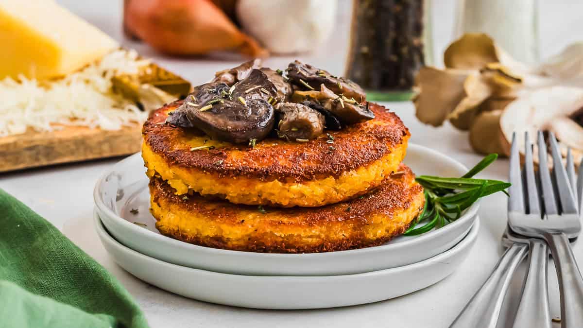 Two golden-brown polenta cakes stacked on a plate, topped with sautéed mushrooms, with herbs and kitchen items in the background.