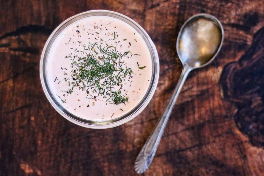 A glass bowl of creamy dip topped with herbs sits next to a metal spoon on a wooden surface.